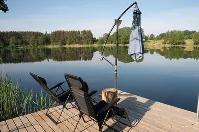 two chairs and an umbrella on a dock next to a lake at Pineplace trio JŪRA in Žaugėdai